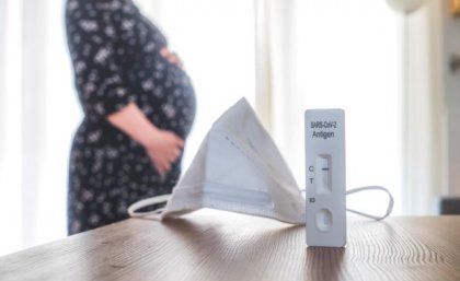 A medical mask and COVID-19 test on desk with pregnant woman in background. Adobe